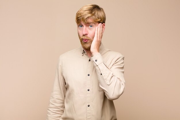 Young blonde man feeling shocked and astonished holding face to hand in disbelief with mouth wide open against flat wall