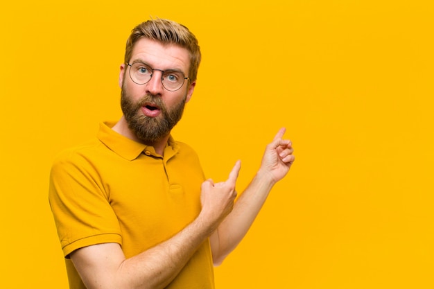 Young blonde man feeling shocked and surprised, pointing to copyspace on the side with amazed, open-mouthed look against orange wall