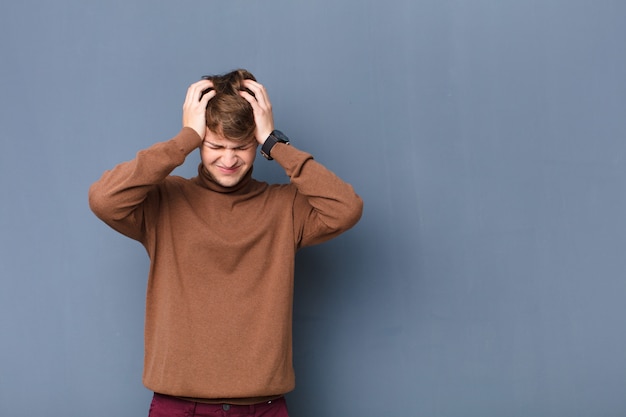 Young blonde man feeling stressed and frustrated, raising hands to head, feeling tired, unhappy and with migraine isolated over wall