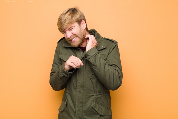 Young blonde man feeling stressed, frustrated and tired, rubbing painful neck, with a worried, troubled look against flat wall