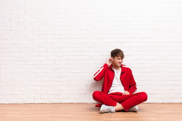 Young blonde man feeling stressed, frustrated and tired, rubbing painful neck, with a worried, troubled look sitting on the floor