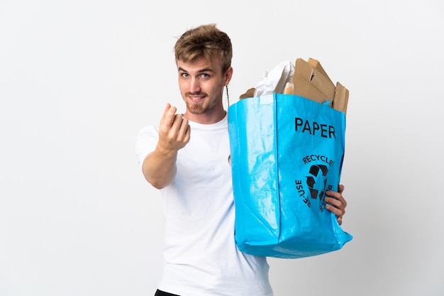 お金のジェスチャーを作る白い背景で隔離のリサイクルする紙でいっぱいのリサイクルバッグを保持している若いブロンドの男