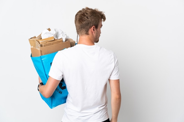 後ろの位置と側面を見て白い壁に隔離されたリサイクルする紙でいっぱいのリサイクルバッグを保持している若いブロンドの男