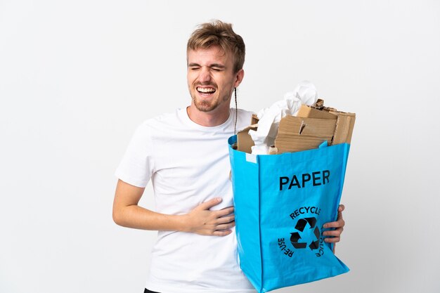 たくさん笑って白い壁に隔離されたリサイクルする紙でいっぱいのリサイクルバッグを保持している若いブロンドの男