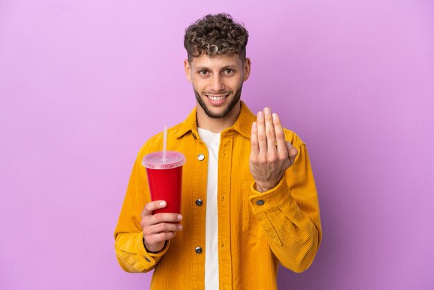 Young blonde man holding soda isolated on purple background inviting to come with hand Happy that you came