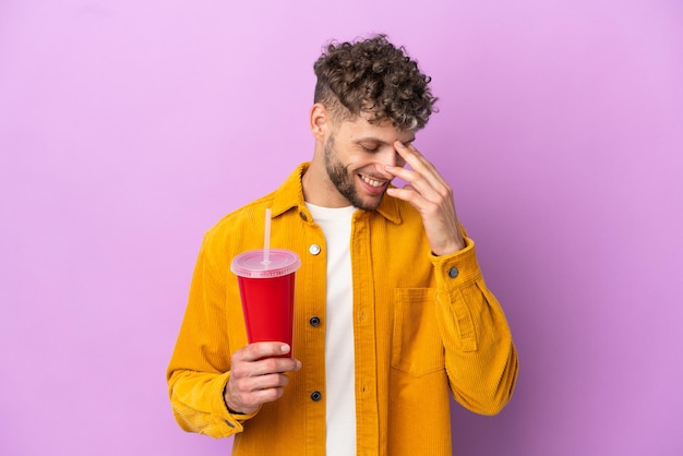 Young blonde man holding soda isolated on purple background laughing