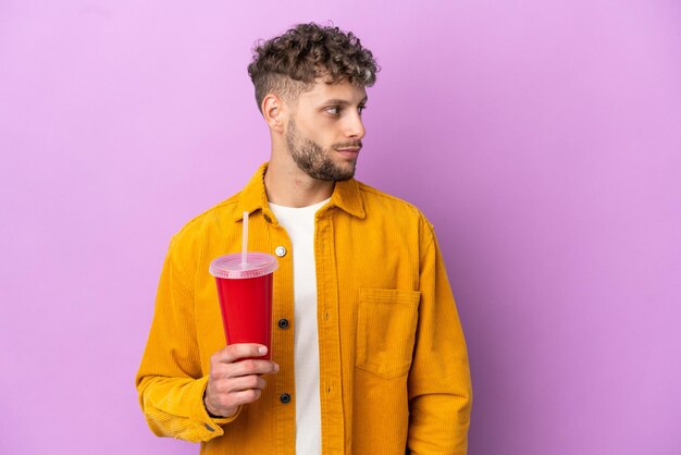 Young blonde man holding soda isolated on purple background looking to the side