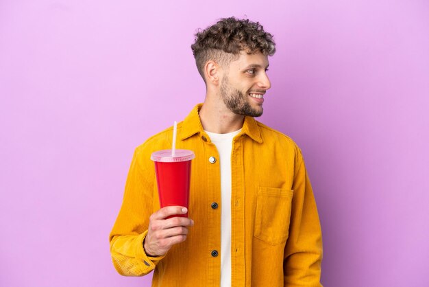 Young blonde man holding soda isolated on purple background looking side