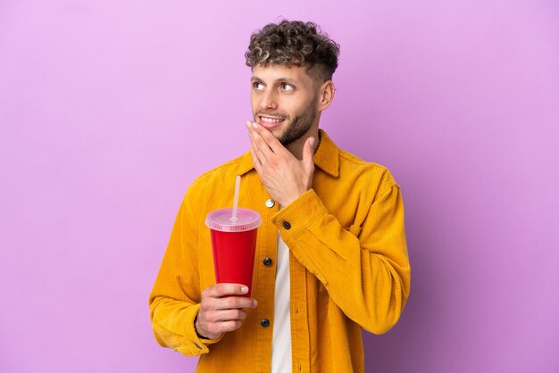 Young blonde man holding soda isolated on purple background looking up while smiling