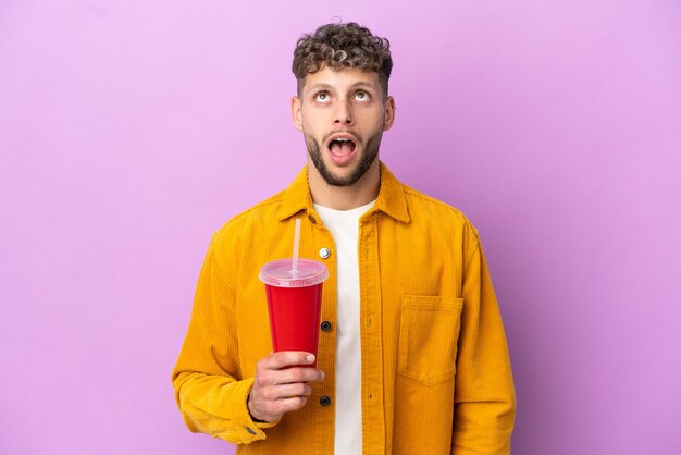 Young blonde man holding soda isolated on purple background looking up and with surprised expression