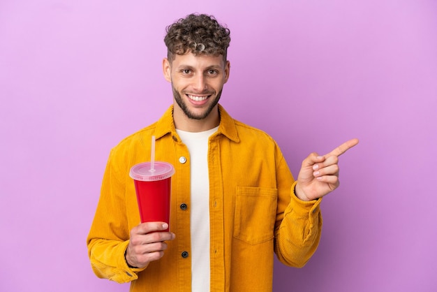 Young blonde man holding soda isolated on purple background pointing finger to the side