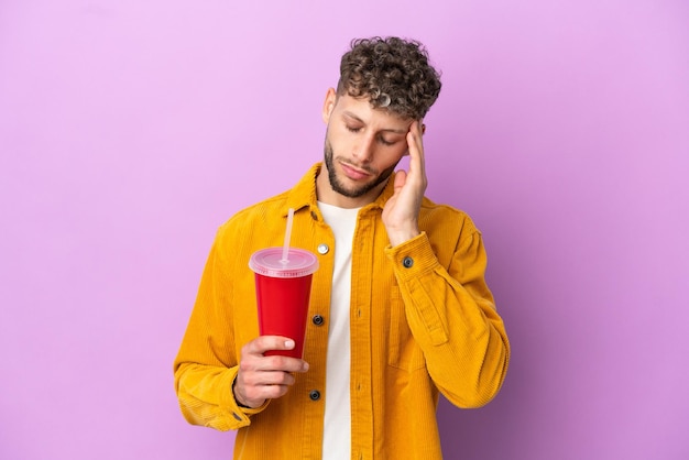 Young blonde man holding soda isolated on purple background with headache