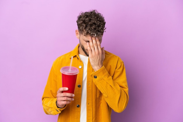 Young blonde man holding soda isolated on purple background with tired and sick expression