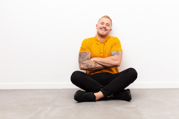 Young blonde man laughing happily with arms crossed, with a relaxed, positive and satisfied pose sitting on cement floor