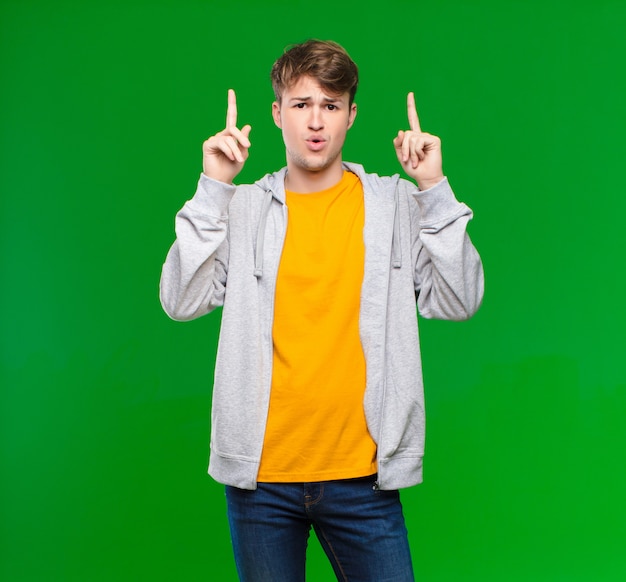 Young blonde man looking shocked, amazed and open mouthed, pointing upwards with both hands over chroma key wall