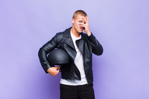 Young blonde man looking shocked, scared or terrified, covering face with hand and peeking between fingers