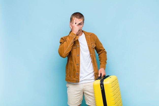 Young blonde man looking shocked, scared or terrified, covering face with hand and peeking between fingers