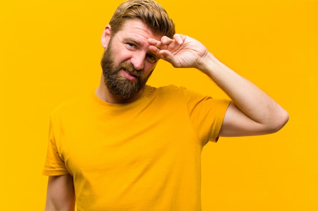 Young blonde man looking stressed, tired and frustrated, drying sweat off forehead, feeling hopeless and exhausted against orange wall