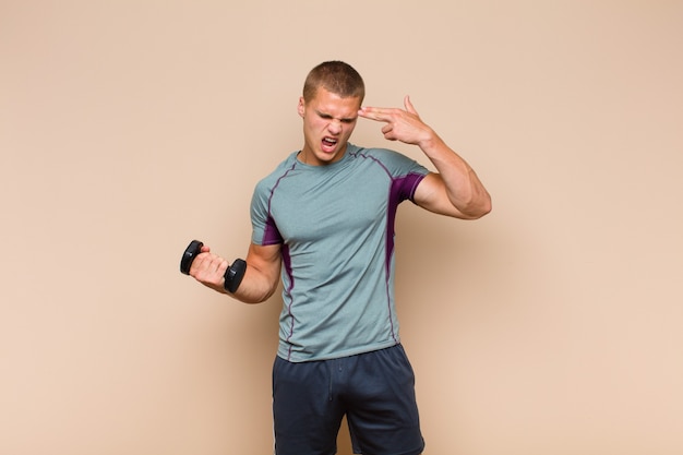 Young blonde man looking unhappy and stressed, suicide gesture making gun sign with hand, pointing to head