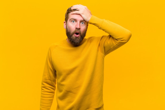 Young blonde man panicking over a forgotten deadline, feeling stressed, having to cover up a mess or mistake on orange wall