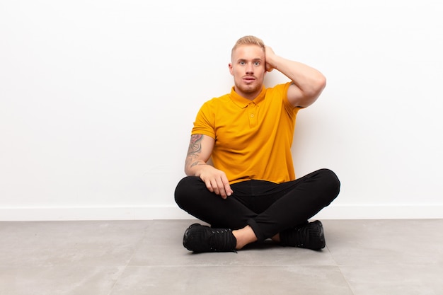 Young blonde man panicking over a forgotten deadline, feeling stressed, having to cover up a mess or mistake sitting on cement floor