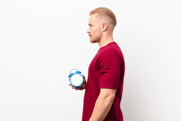 Young blonde man on profile view looking to copy space ahead, thinking, imagining or daydreaming on white wall holding an alarm clock