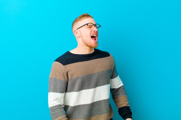 Young blonde man screaming furiously, shouting aggressively, looking stressed and angry against blue wall