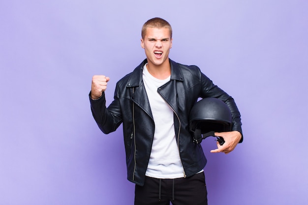 Young blonde man shouting aggressively with an angry expression or with fists clenched celebrating success