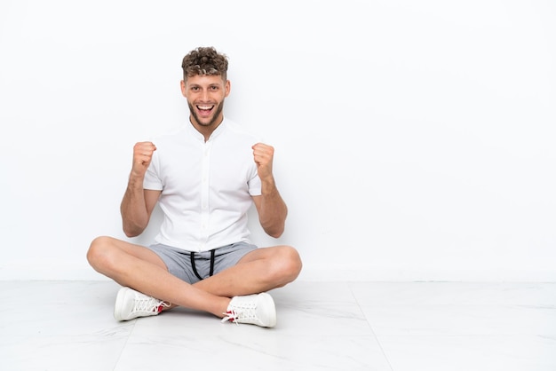 勝者の位置での勝利を祝って白い背景で隔離の床に座っている若いブロンドの男