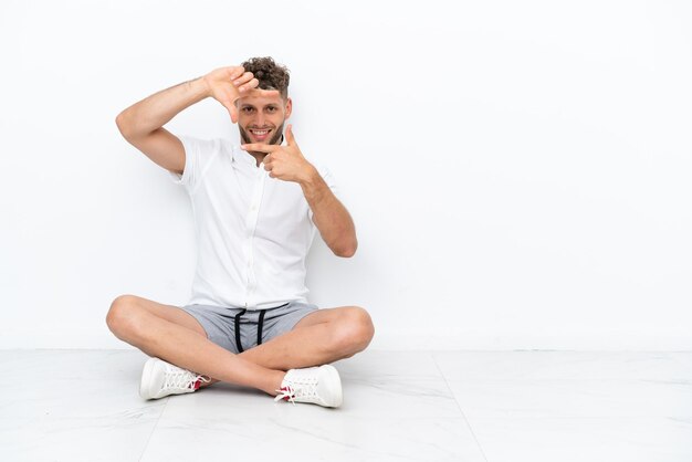 Photo young blonde man sitting on the floor isolated on white background focusing face. framing symbol