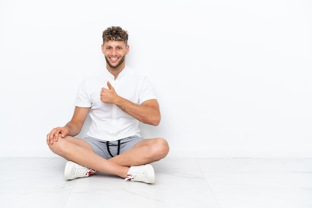 Young blonde man sitting on the floor isolated on white background giving a thumbs up gesture