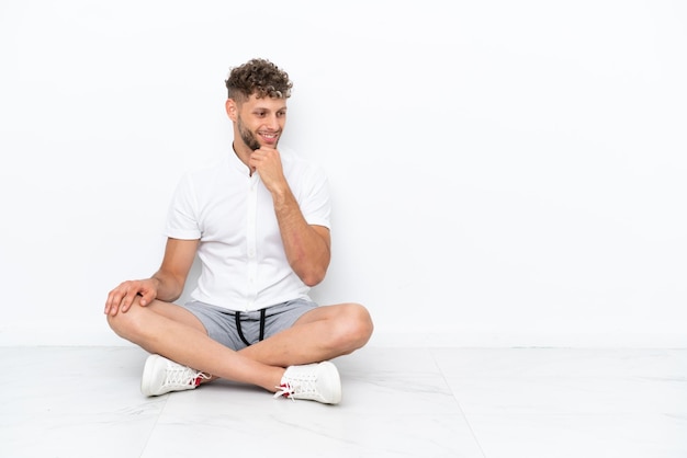 Foto giovane biondo seduto sul pavimento isolato su sfondo bianco guardando al lato