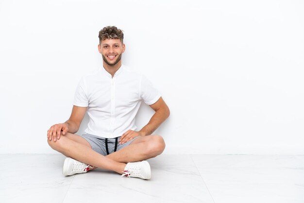 Young blonde man sitting on the floor isolated on white background posing with arms at hip and smiling