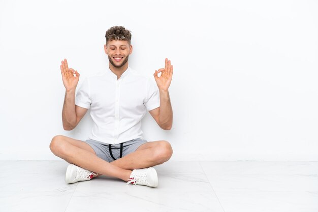 Young blonde man sitting on the floor isolated on white background in zen pose