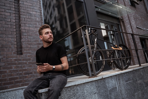 Giovane biondo seduto sul parapetto del muro di pietra vicino alla sua bici da strada mentre messaggia, mi piace, posta sui social media guardando pensieroso a distanza indossando jeans neri e t-shirt