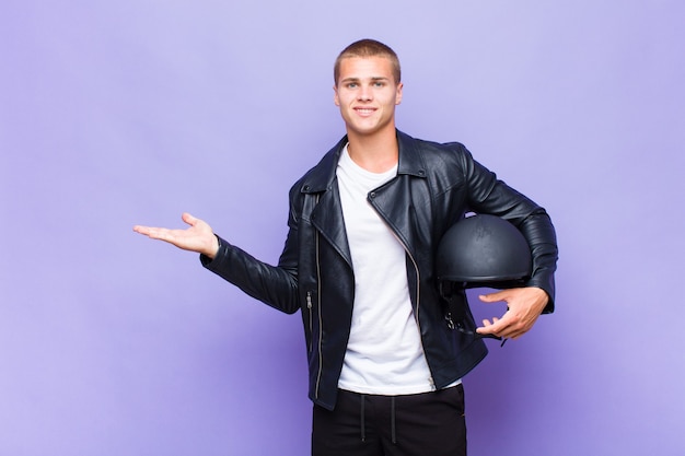 Young blonde man smiling cheerfully, feeling happy and showing a concept in copy space with palm of hand