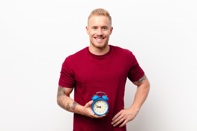 Young blonde man smiling happily with a hand on hip and confident, positive, proud and friendly attitude against white wall holding an alarm clock