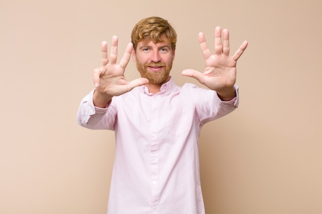 Young blonde man smiling and looking friendly, showing number nine or ninth with hand forward, counting down against flat wall