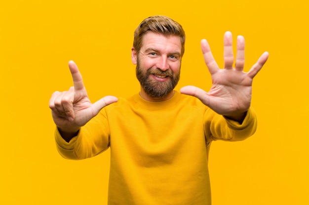 Young blonde man smiling and looking friendly, showing number sevenseventh with hand forward, counting down  orange wall