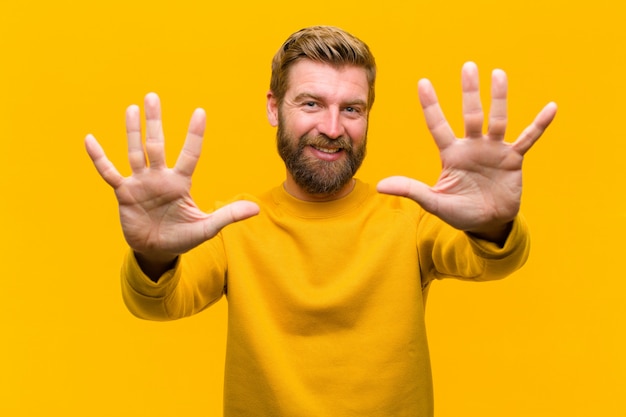 Foto giovane uomo biondo che sorride e che sembra amichevole, mostrando numero dieci o decimo con la mano in avanti, conto alla rovescia contro la parete arancione
