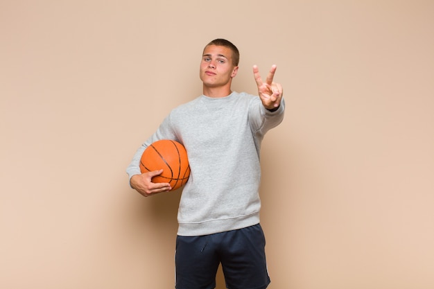 Young blonde man smiling and looking happy, carefree and\
positive, gesturing victory or peace with one hand