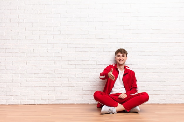 Young blonde man smiling, looking happy, confident and friendly, offering a handshake to close a deal, cooperating sitting on the floor