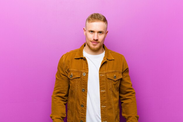 Young blonde man smiling positively and confidently, looking satisfied, friendly and happy against purple wall