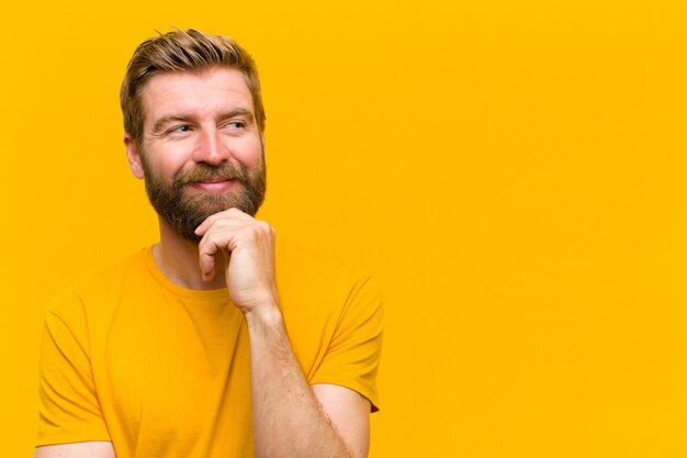 Young blonde man smiling with a happy, confident expression with hand on chin, wondering and looking to the side against orange wall