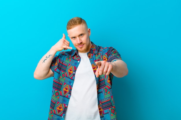 Young blonde man wearing a print shirt smiling cheerfully and pointing to camera while making a call you later gesture, talking on phone