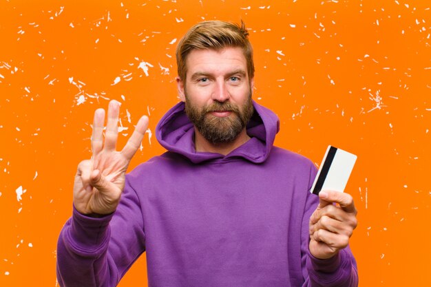 Young blonde man with a credit card  wearing a purple hoodie against damaged orange wall