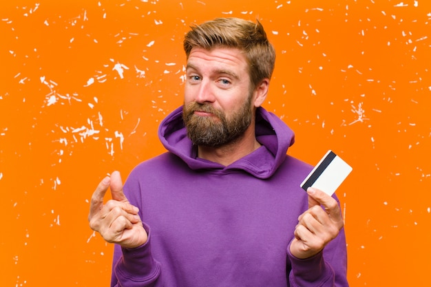 Foto giovane uomo biondo con una carta di credito che indossa una felpa con cappuccio viola contro la parete arancione danneggiata