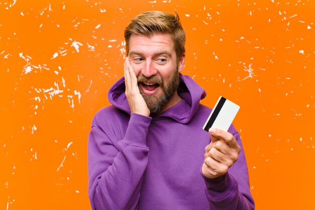 Foto giovane uomo biondo con una carta di credito che indossa una felpa con cappuccio viola contro la parete arancione danneggiata