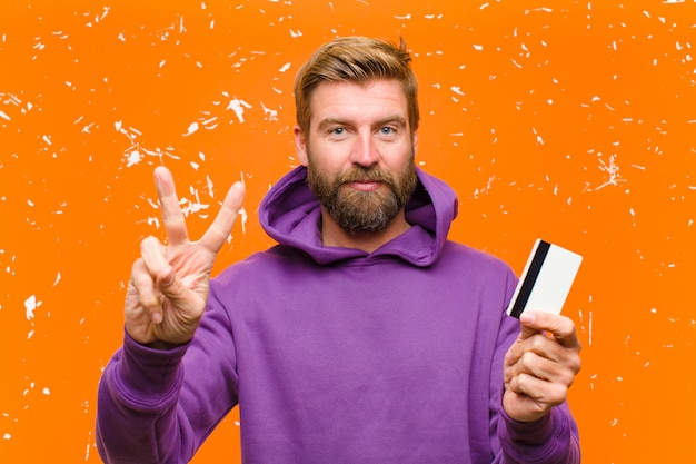 Young blonde man with a credit card  wearing a purple hoodie against damaged orange wall