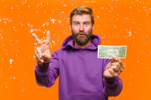 Young blonde man with dollar bills or banknotes wearing a purple hoodie against damaged orange 